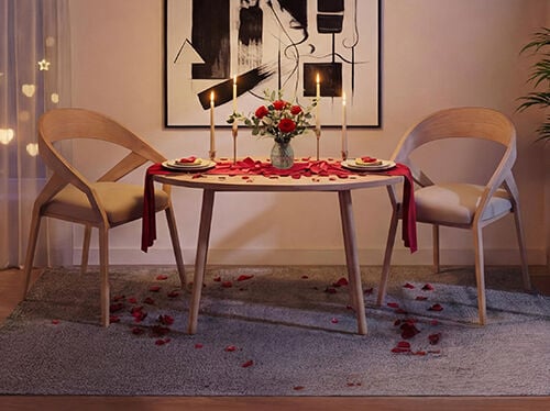 Romantic dining table set for two in a cozy living room, featuring a round wooden table with matching modern chairs, soft candlelight, a vase of red roses, scattered rose petals on the table and floor, and warm ambient lighting creating an intimate Valentine&rsquo;s Day atmosphere.