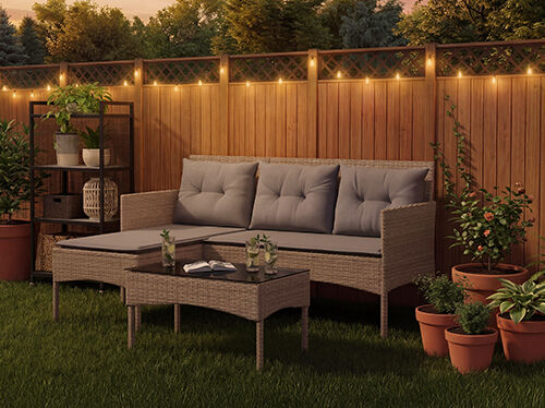 Rattan garden lounge set with grey cushions including a three-seat sofa, footstool and coffee table on a lawn with string lights and potted plants