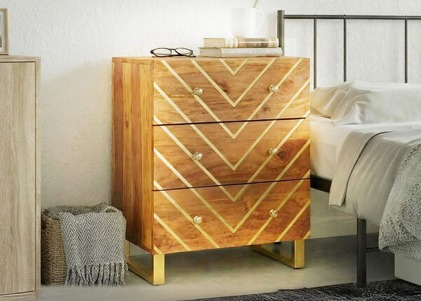 Wooden bedside table with three drawers and a geometric gold pattern, placed next to a bed in a bright bedroom.