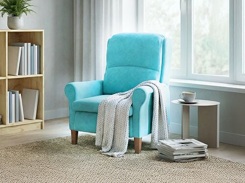 A turquoise armchair next to a window, with a throw blanket, side table, and stacked books in a bright, cosy living space.