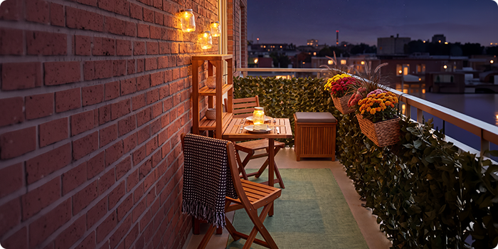 Small balcony with wooden chairs and table, illuminated by lanterns and candles, decorated with potted flowers and greenery, overlooking a city skyline at night.
