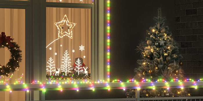 An outdoor Christmas scene featuring a decorated tree with golden ornaments and lights next to a window framed by multicolored string lights. The window displays illuminated decorations with stars, snowflakes, and tree shapes, and a wreath with a red bow hangs on the adjacent window.