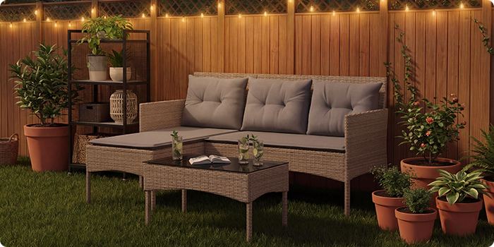 Rattan garden lounge set with grey cushions including a three-seat sofa, footstool and coffee table on a lawn with string lights and potted plants