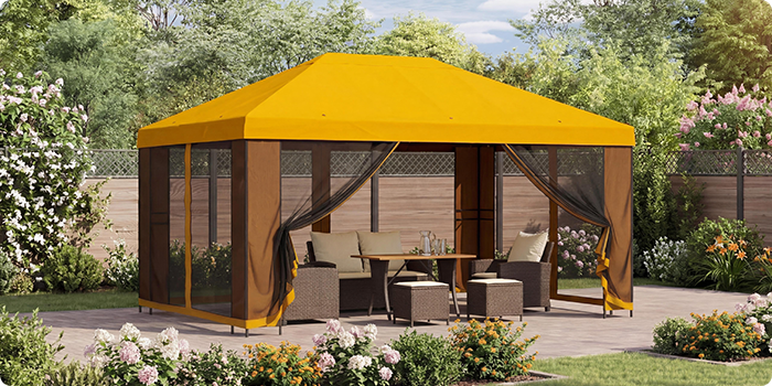 Yellow garden gazebo with mesh curtains, sheltering a lounge seating set with a table on a patio, bordered by blooming plants in a sunny garden setting. 